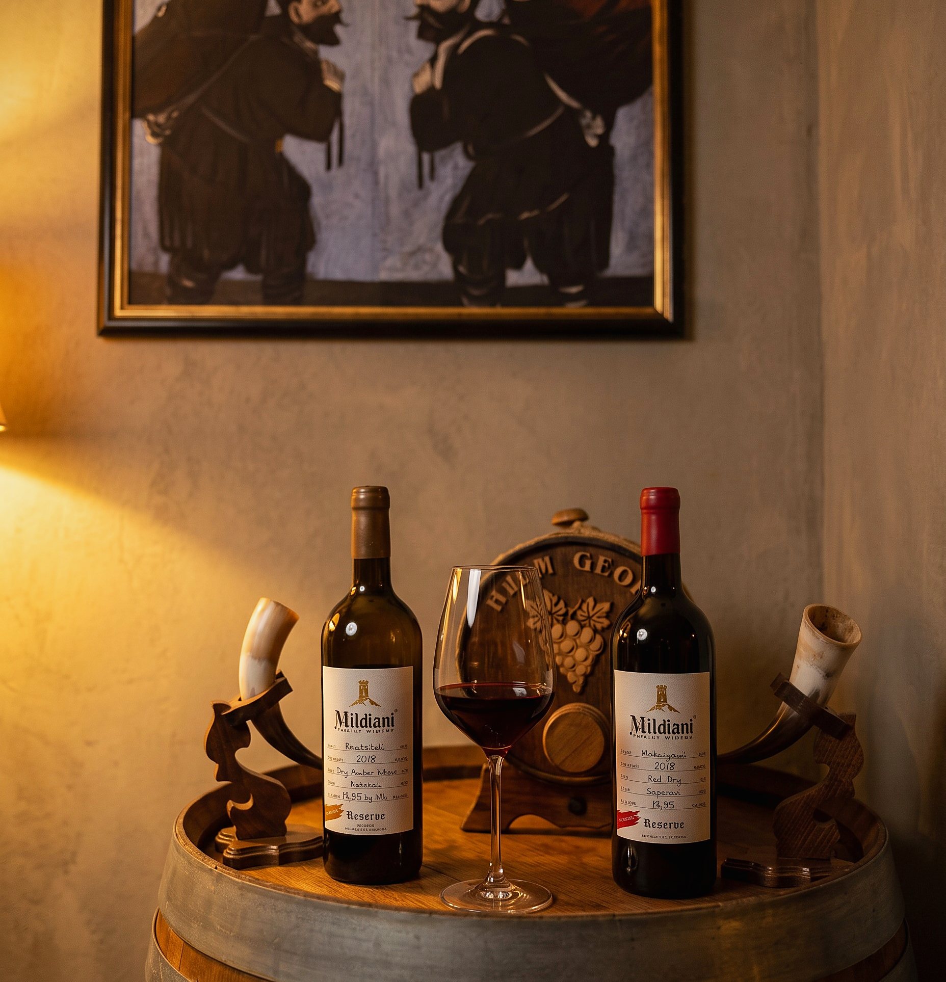 Zwei Flaschen Mildiani Reserve, ein eingeschenktes Glas und zwei Trinkhörner auf dem Eichenfass, darüber ein georgisches Gemälde